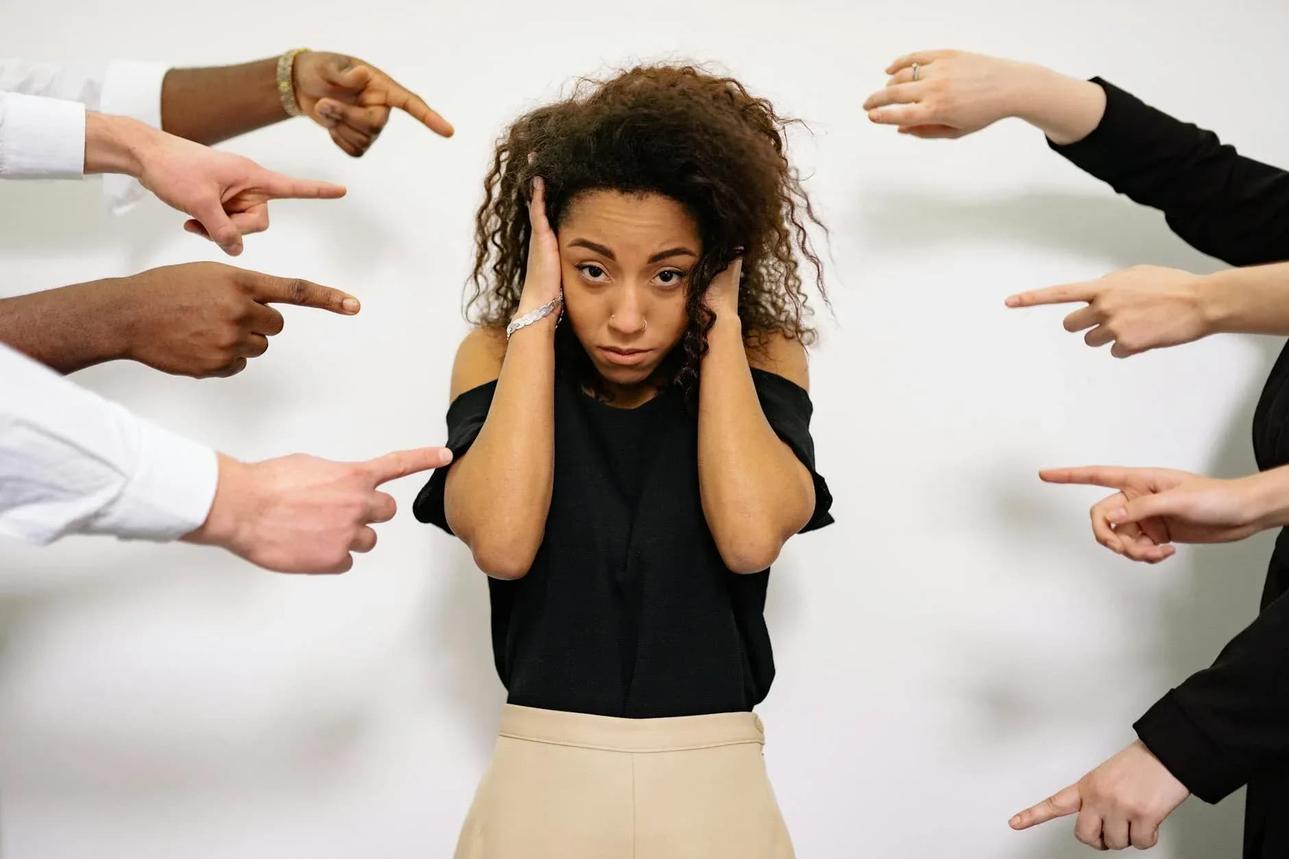 A stressed woman with hands on ears surrounded by pointing fingers, symbolizing pressure and stress.