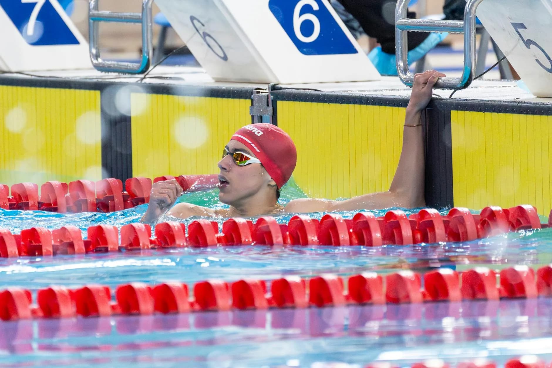 A swimmer in a red cap and goggles celebrates victory after finishing a race, holding the lane divider.