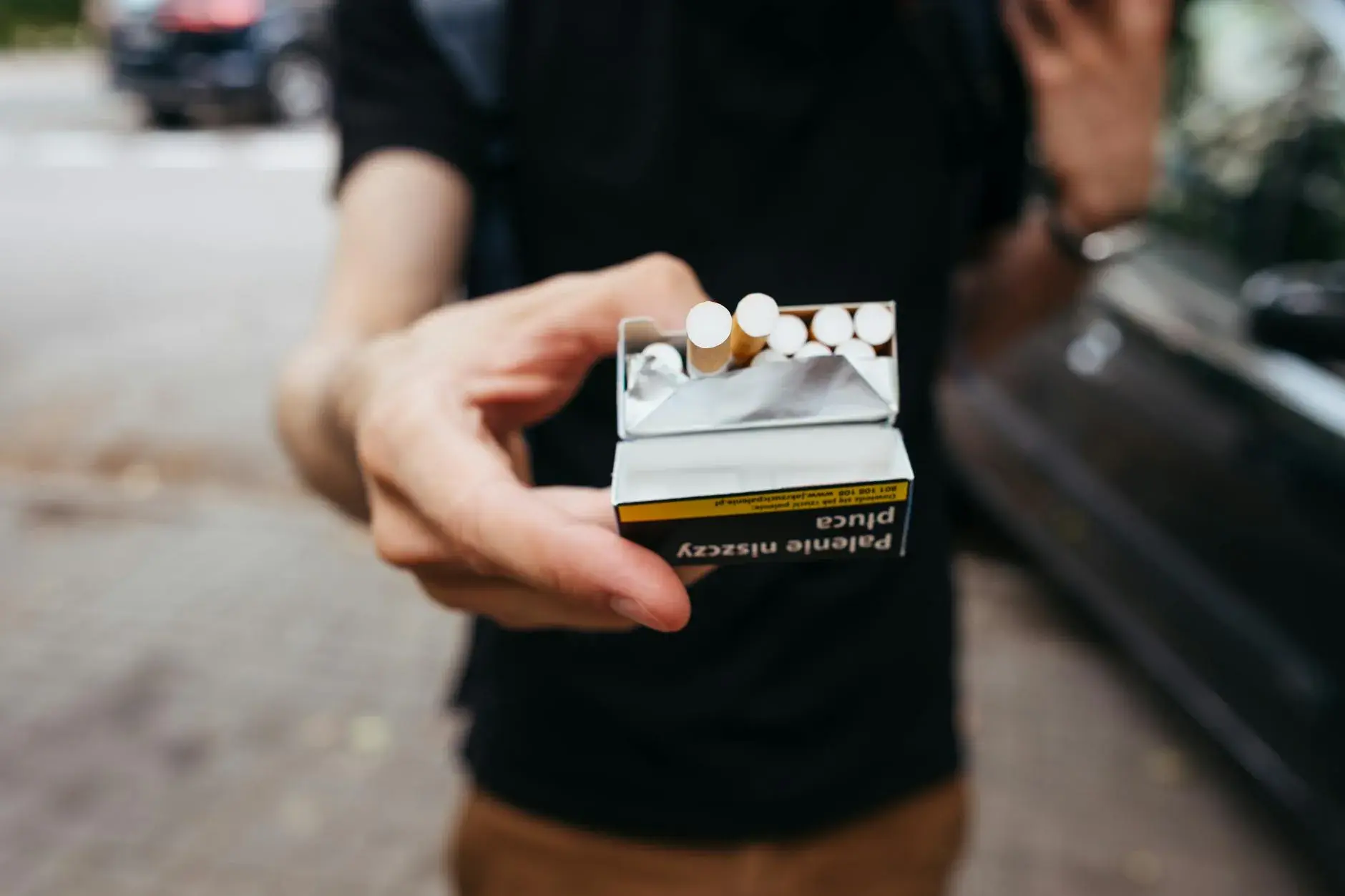 Et close-up af en mand, der rækker en pakke cigaretter frem udendørs på en bygade. Stemningen er hverdagsnær og dokumentarisk.