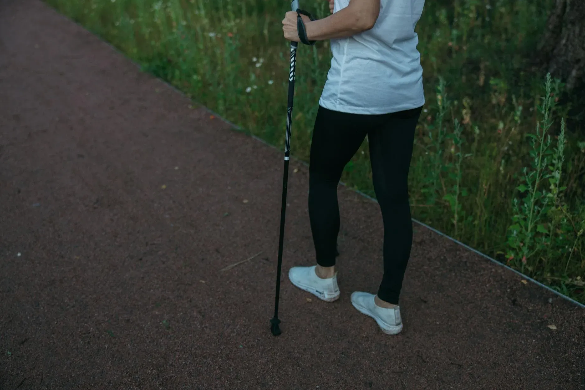 En person i sportstøj går på en skovsti og bruger vandrestav for stabilitet og fremdrift. Billedet formidler friluftsliv og fysisk aktivitet.