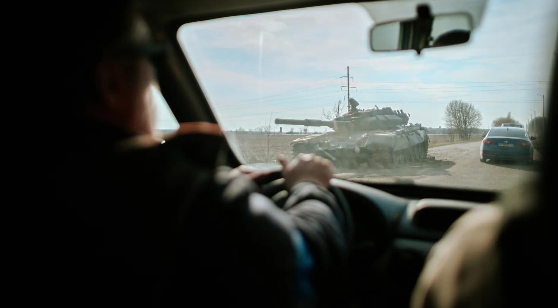 Et billede taget fra en bil, der passerer en militær tank på en vej i Ukraine. Scenen viser en almindelig vej med en militær tilstedeværelse.