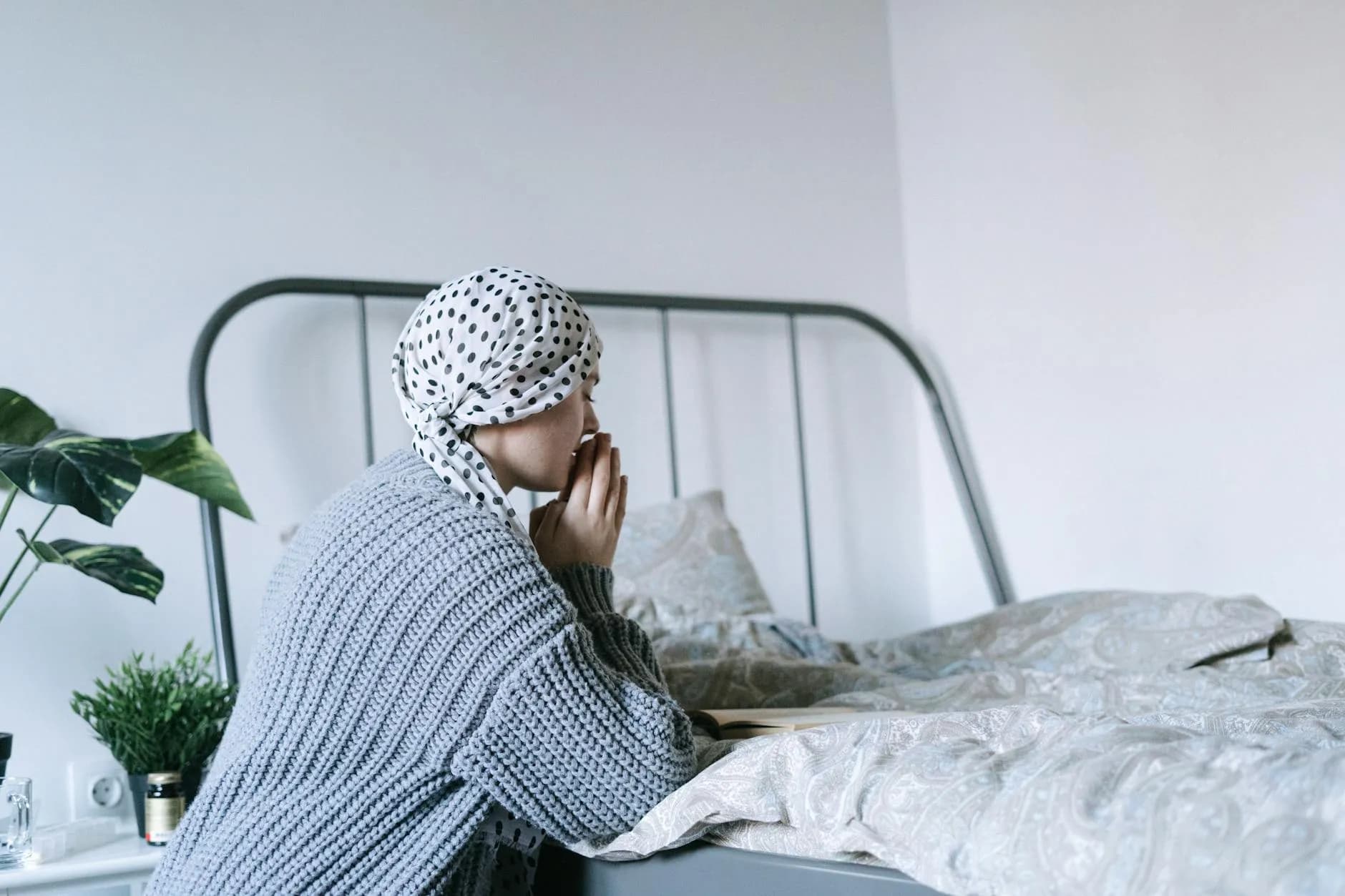 A woman in a headscarf sits by her bed, lost in thought as she gathers strength for what lies ahead.
