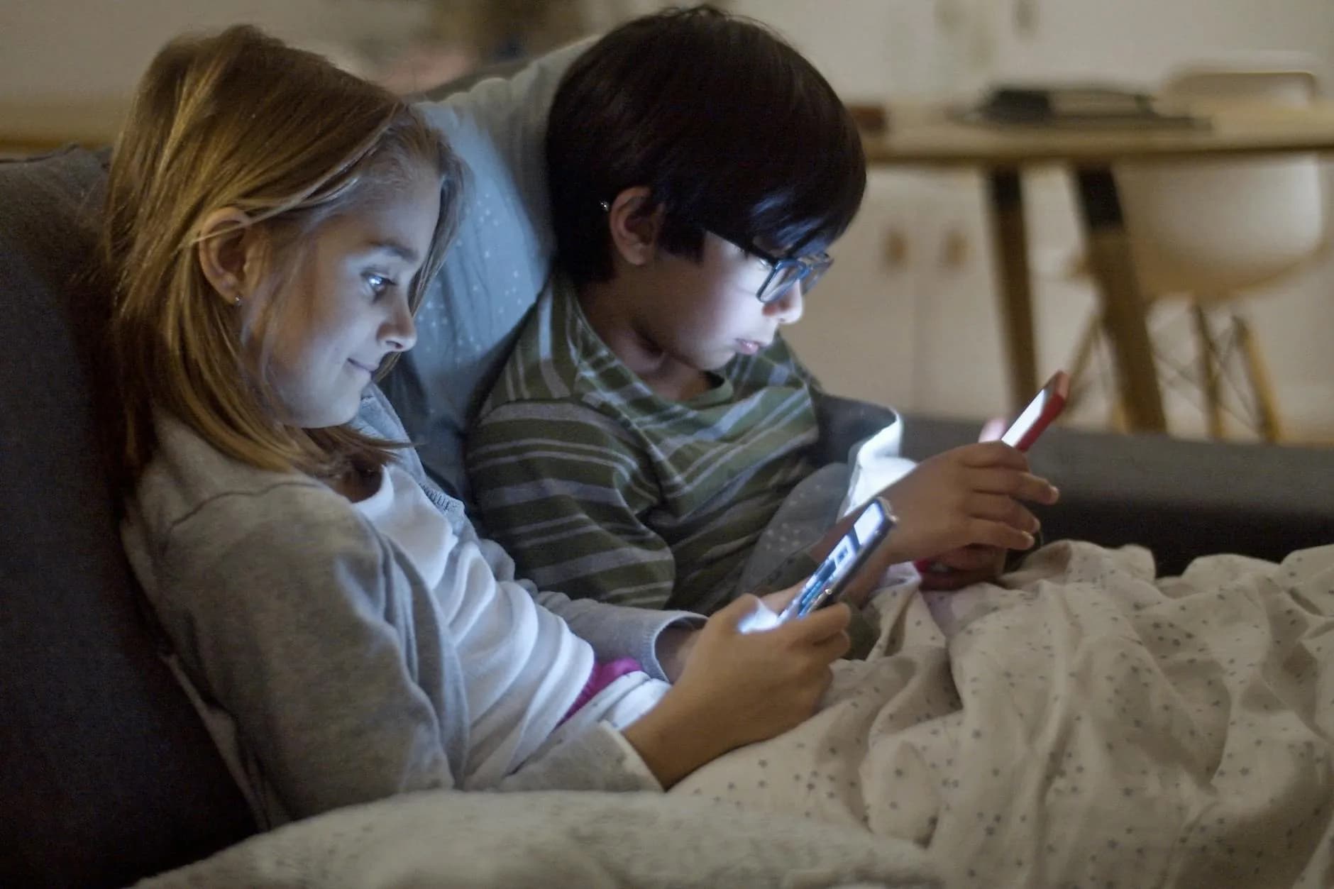 To børn sidder indendørs og bruger smartphones om natten, gemt under tæpper. Billedet illustrerer skærmtid i en privat og mørk setting.
