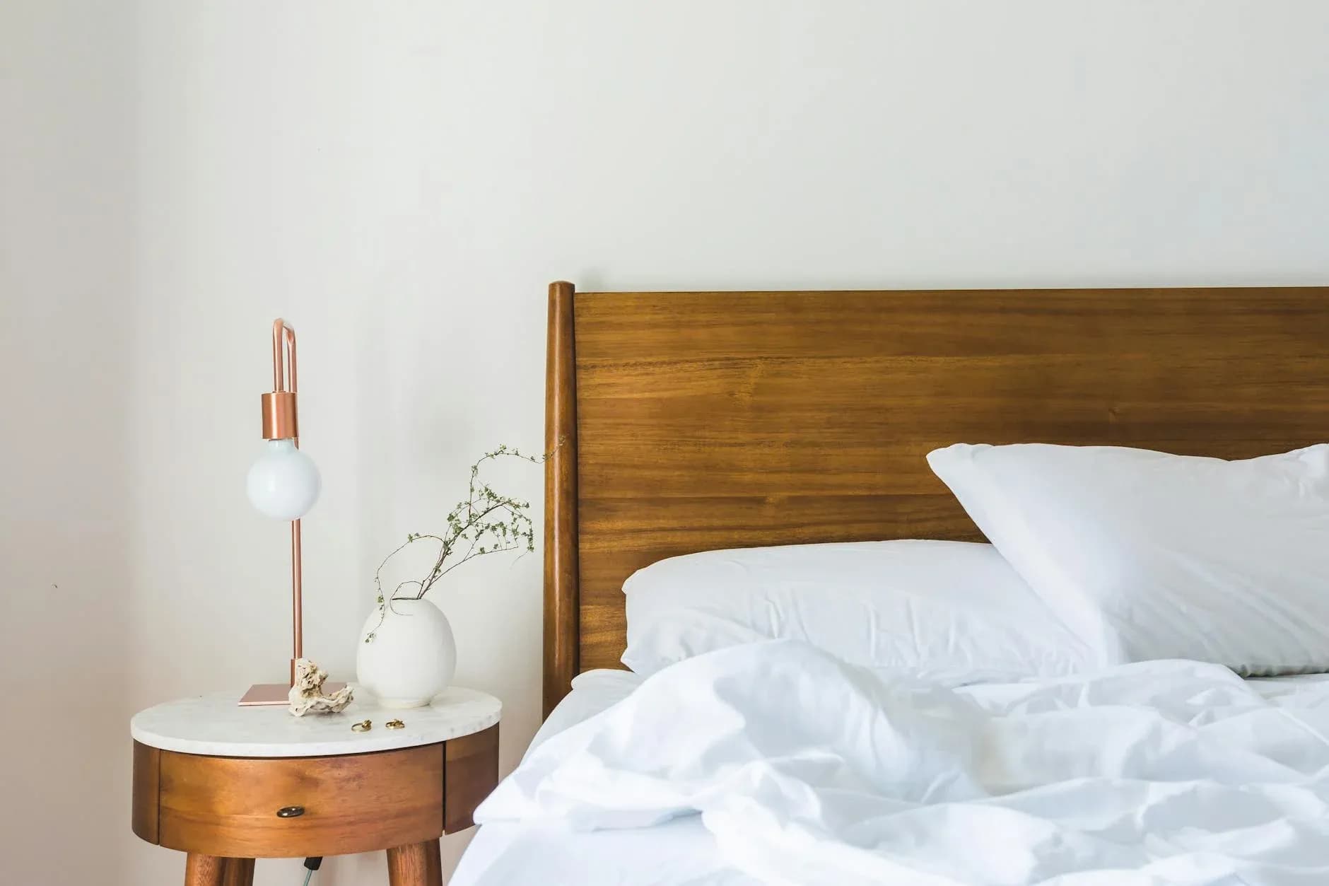A serene minimalist bedroom showcases a natural wooden headboard, crisp white bedding, and a modern side table lamp casting warm light across the space.