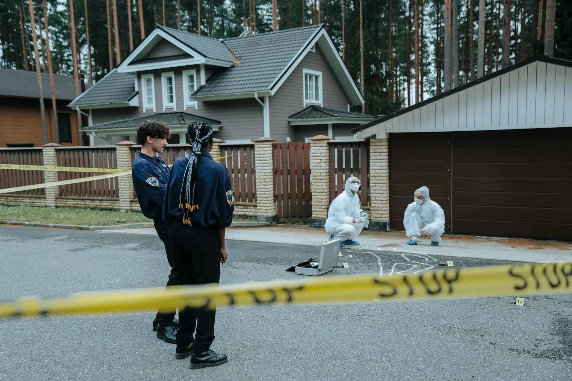 Politi og retsmedicinske eksperter undersøger et gerningssted foran forstædernes huse. Scenen formidler efterforskning og dokumentation.