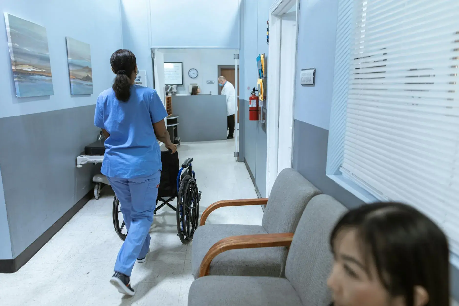 En sygeplejerske i scrubs skubber en kørestol gennem en veloplyst hospitalsgang. Scenen illustrerer patientpleje og omsorg i sundhedsvæsenet.