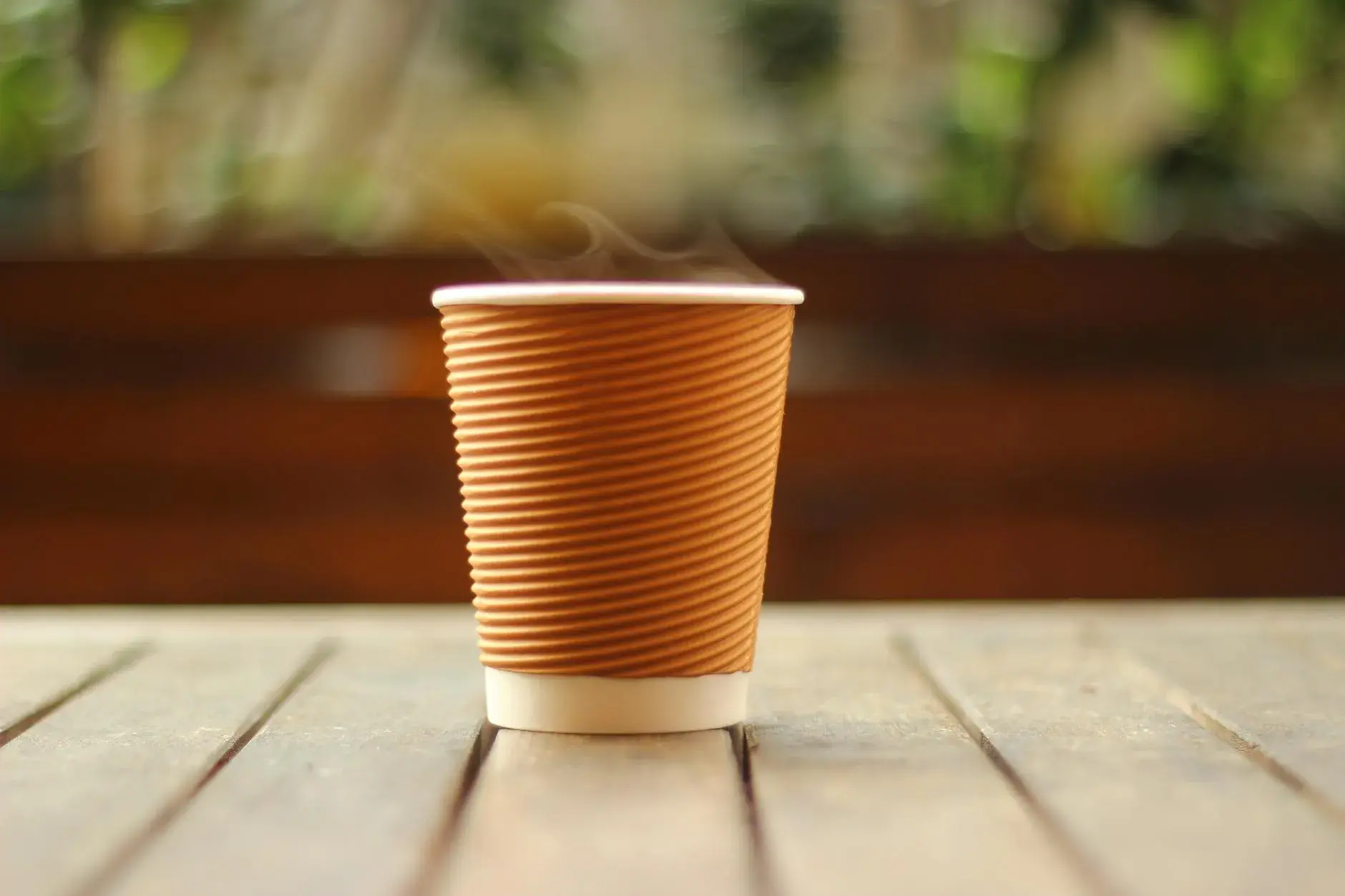 Et nærbillede af en dampende kop kaffe i et brunt papkrus på et træbord indendørs. Varmen og dampen skaber en hyggelig stemning.