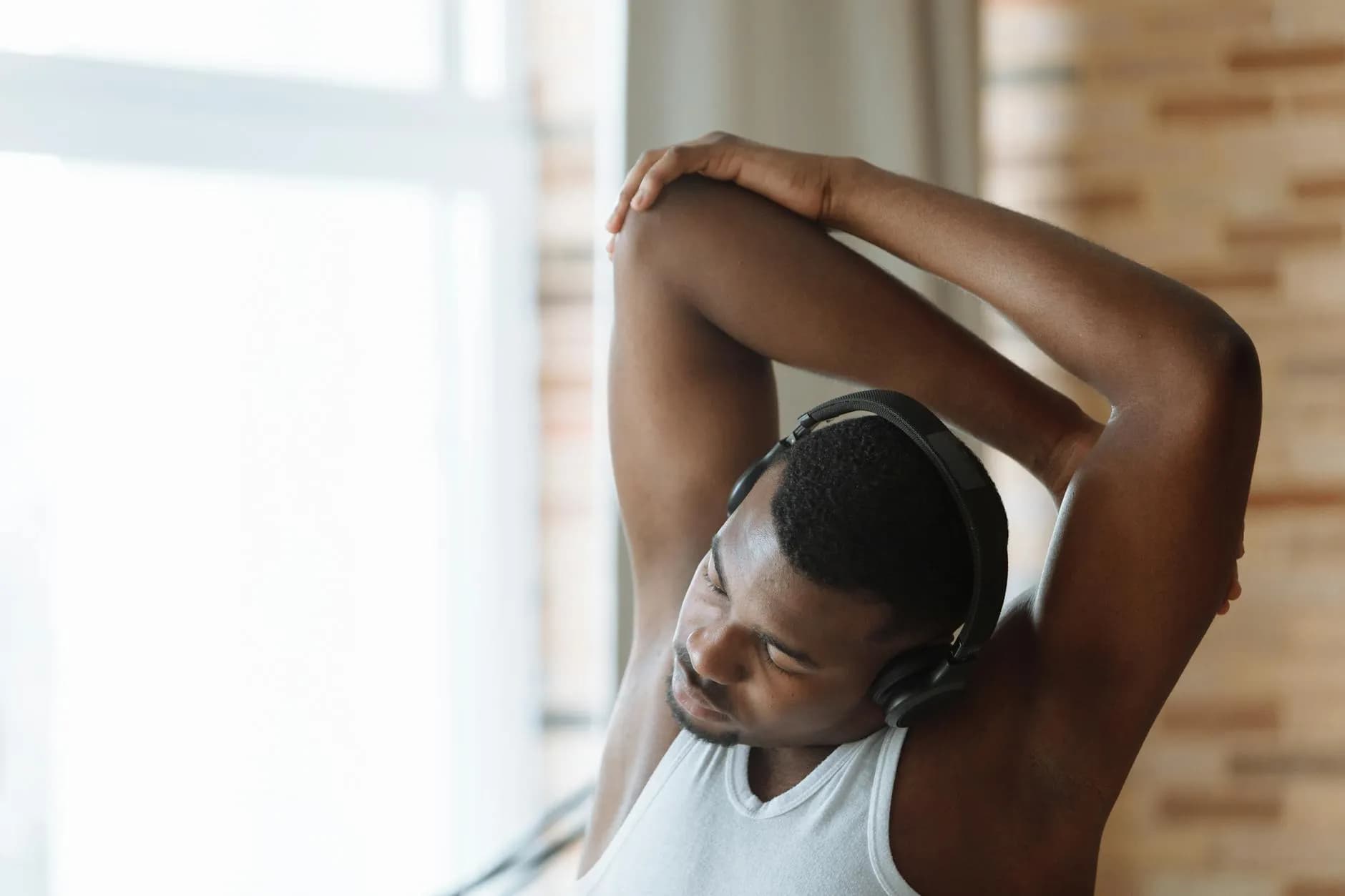 En mand i hvid tanktop strækker ud indendørs, mens han lytter til musik via høretelefoner. Billedet formidler ro, fokus og opvarmning før træning.