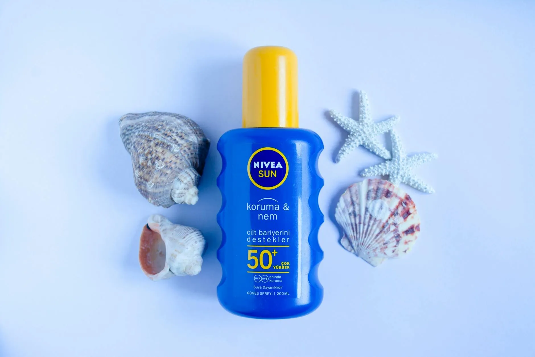 En blå Nivea Sun SPF 50+ solcremeflaske står på en hvid baggrund sammen med havskaller og stjerneformede dekorationer. Motivet understreger sommerhudpleje og solbeskyttelse.