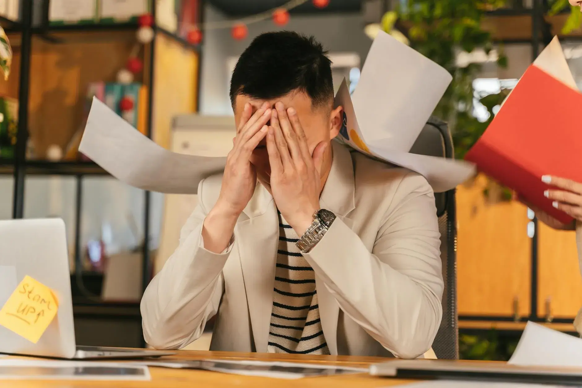 En stresset forretningsmand sidder i et kontormiljø omgivet af papirarbejde, der illustrerer arbejdsrelateret udmattelse. Billedet formidler tydeligt temaet burnout og pres i hverdagen.