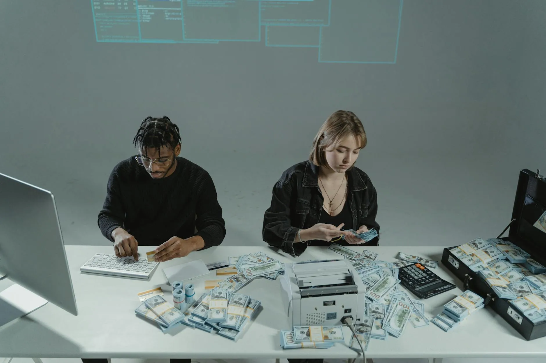 To personer tæller penge ved et skrivebord med computere og kontantudstyr. Billedet formidler arbejde med økonomi, betalinger eller kassehåndtering.
