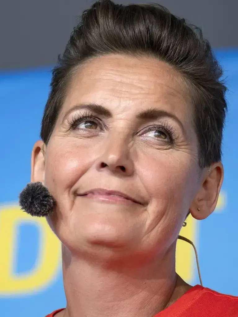 En kvindelig politiker holder en tale, mens bodyguards står i baggrunden, og et amerikansk flag ses bagved. Billedet formidler politisk kommunikation og offentlig fremtræden.