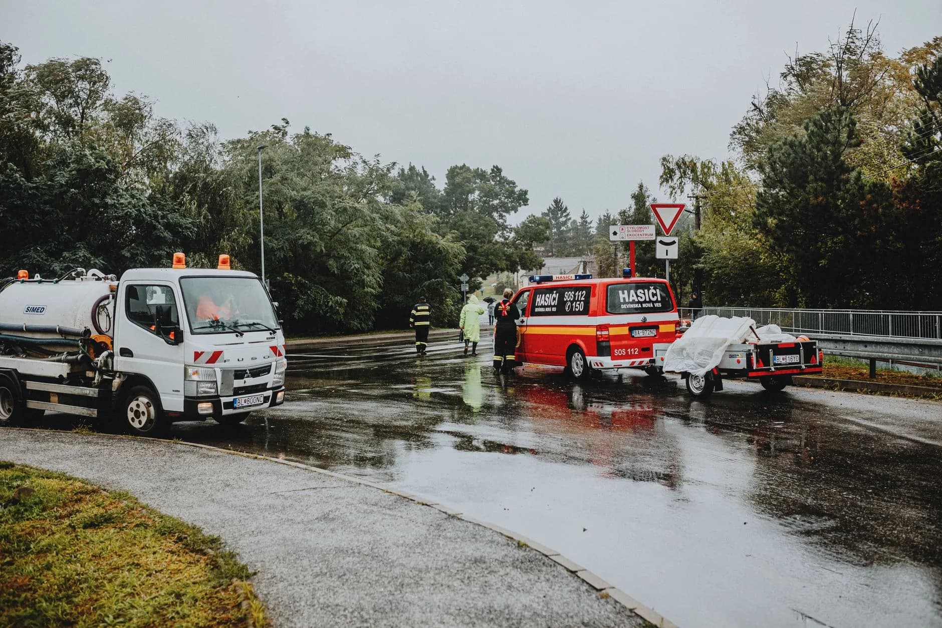 Redningspersonale og køretøjer er samlet ved en hændelse på en regnvåd vej. Scenen er fotograferet i dårligt vejr med synlige redningsindsatser.