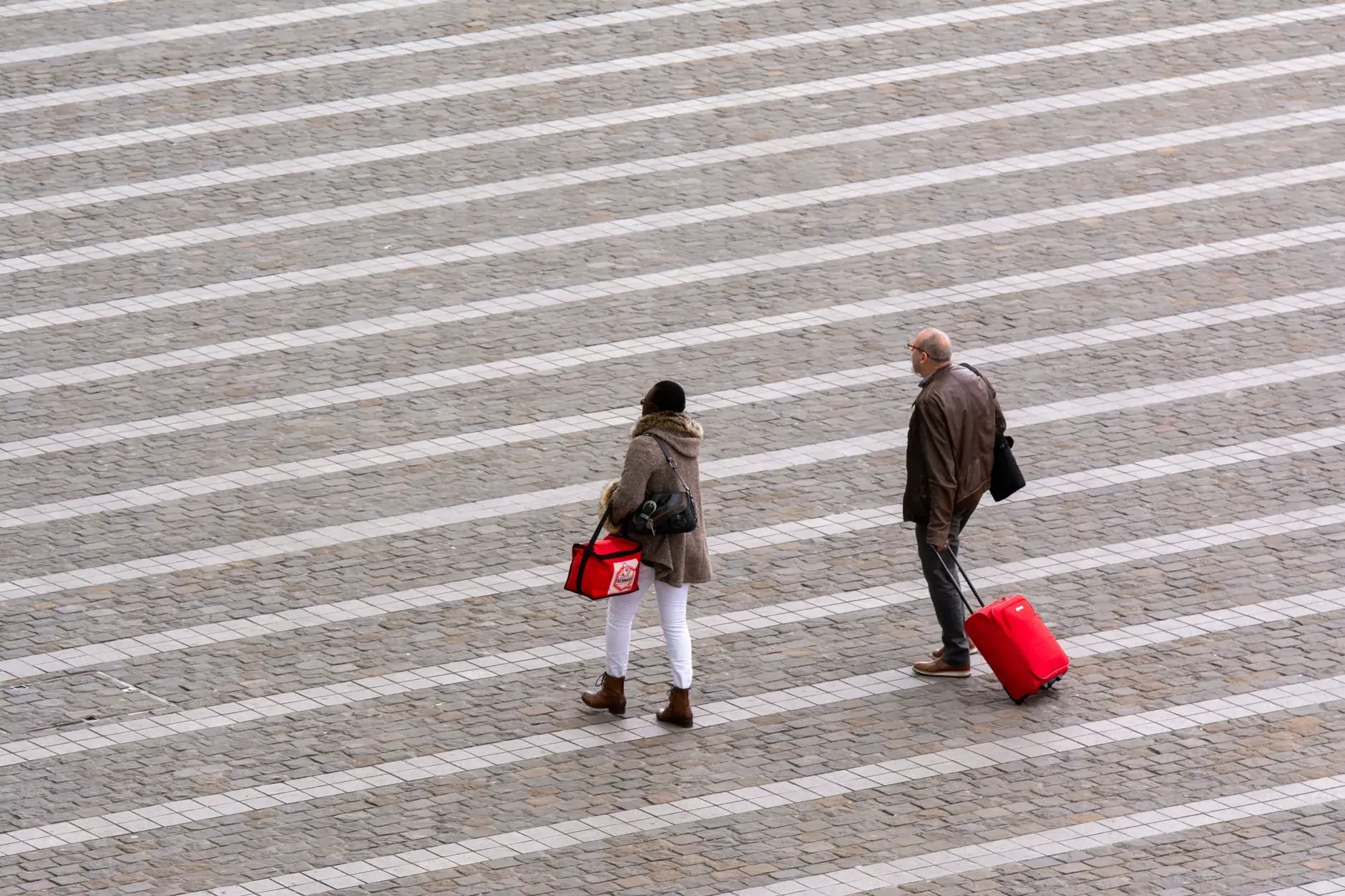 Et aerialt udsnit af mennesker, der går med kufferter hen over en mønstret bybelægning. Motivet understreger urban rejse og bevægelse.