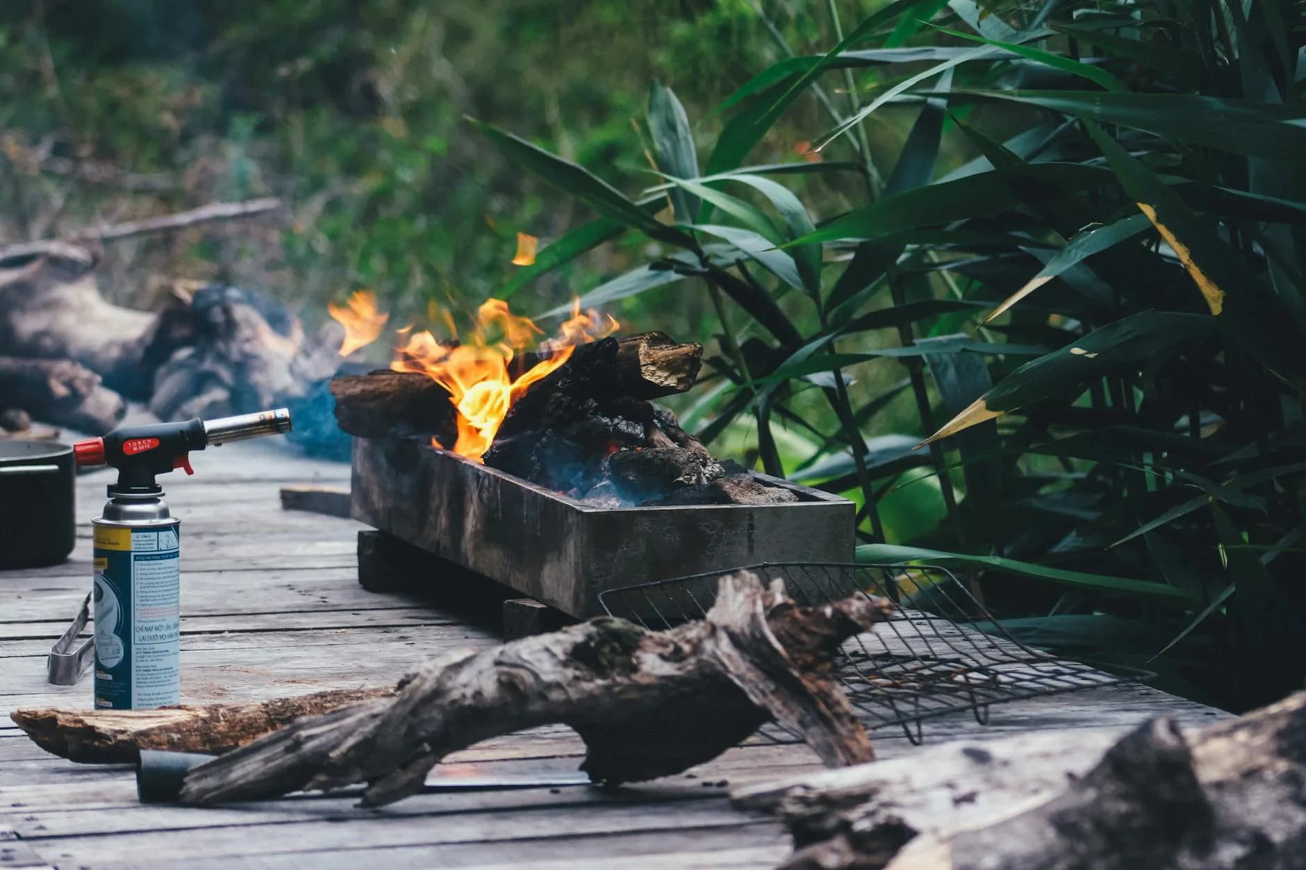 En rustik udendørs grillopsætning med brændestykker og synlige flammer i frodig, grøn beplantning. Stemningen er varm og rå, som taget fra en hyggelig haveaften.