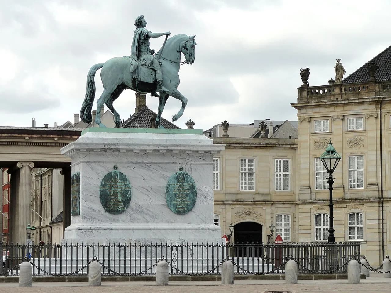 En statue af Frederik V står i gården ved Amalienborg Palace i København. Billedet fremhæver monumentet i et klassisk, arkitektonisk miljø.