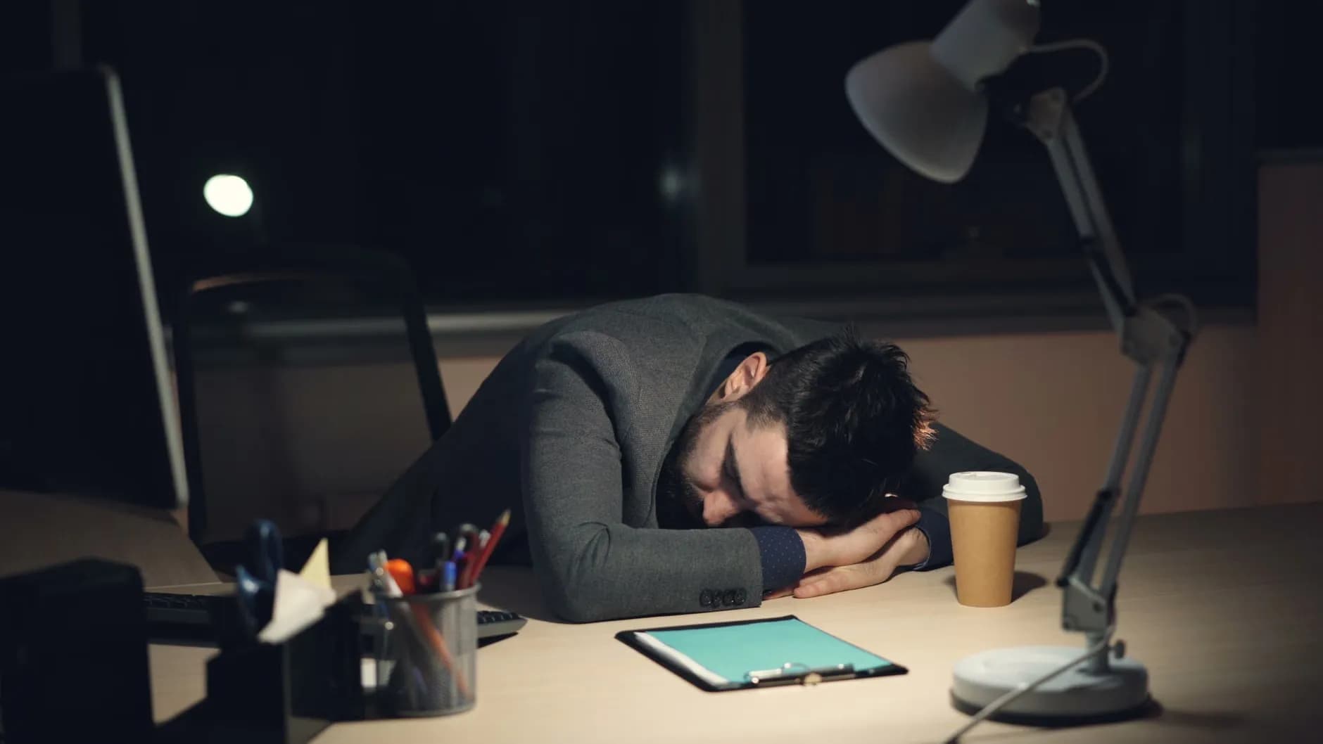En mand i jakkesæt sover på skrivebordet med en kop kaffe og en tablet ved siden af, symboliserende træthed på arbejdspladsen.