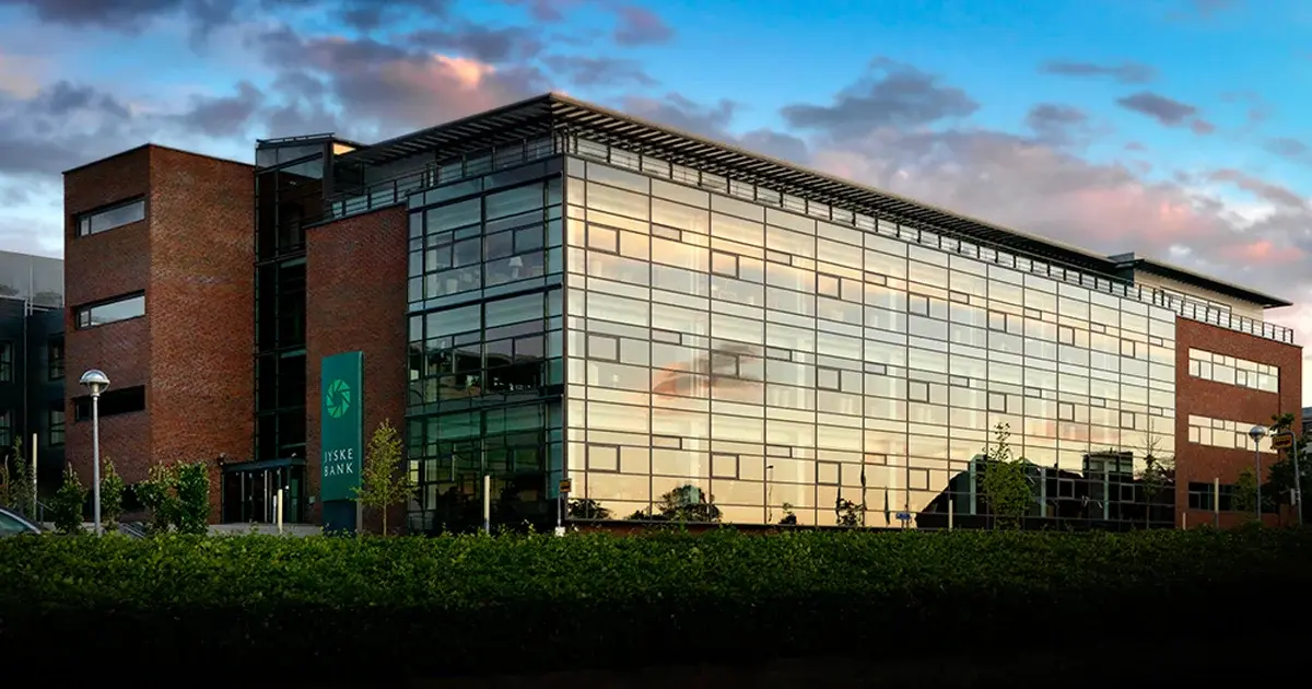 Modern Jyske Bank headquarters building with glass and brick facade reflecting a sunset sky.