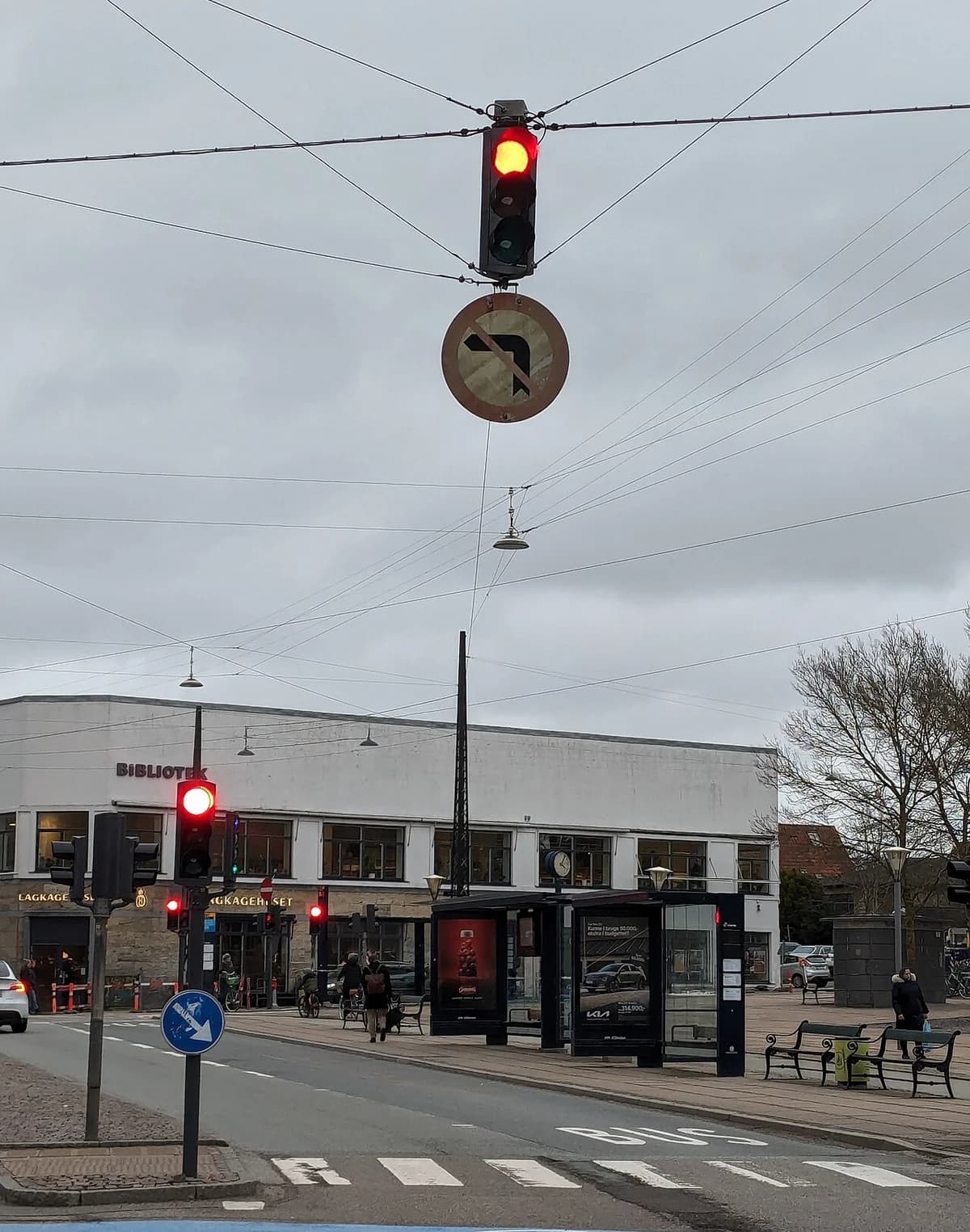A city intersection with red traffic lights and an old no-left-turn sign hanging below one light.