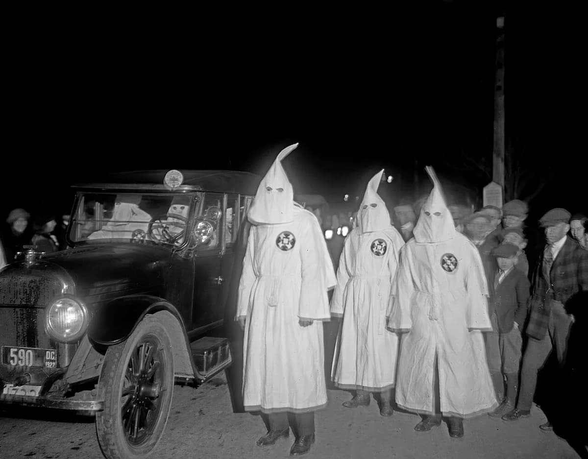Three Ku Klux Klan members in robes stand beside a car at night during a parade.