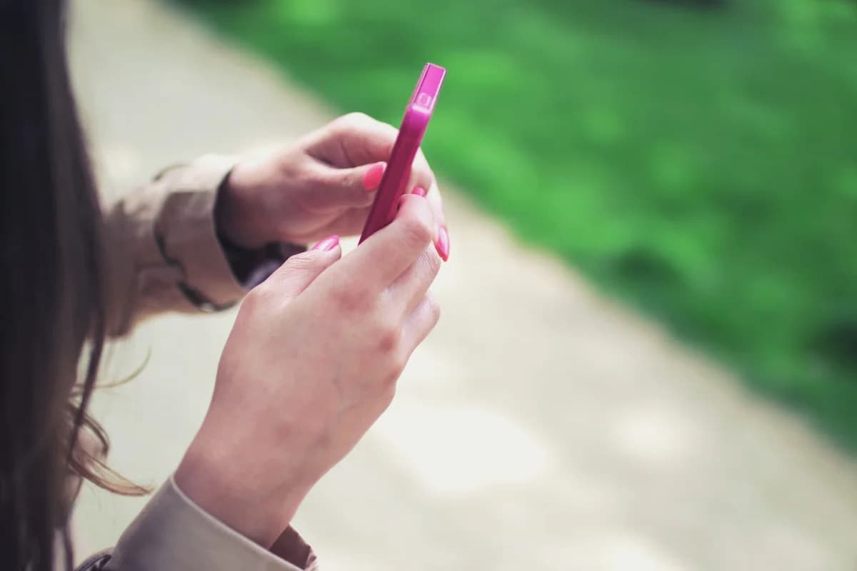 A woman holding a pink smartphone outdoors near a path.