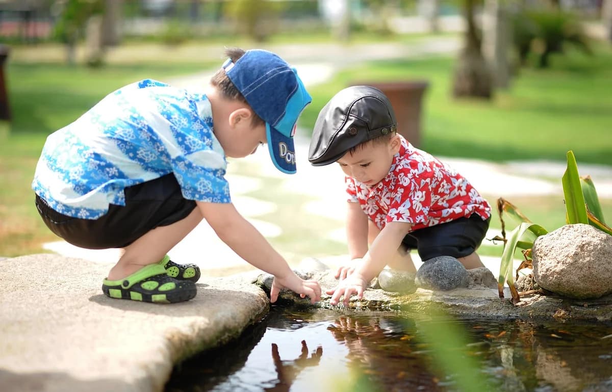 Two young boys crouch by a pond and play in a park.
