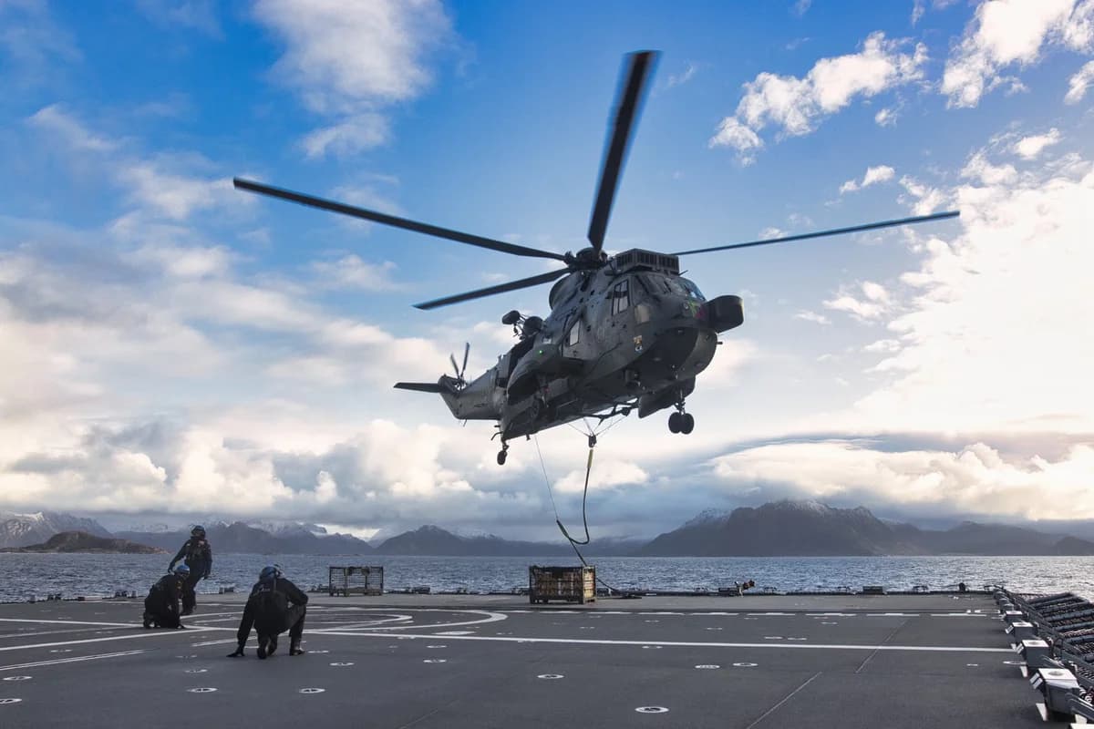 A military helicopter hovers above a ship deck with three crew members nearby and mountains in the distance.