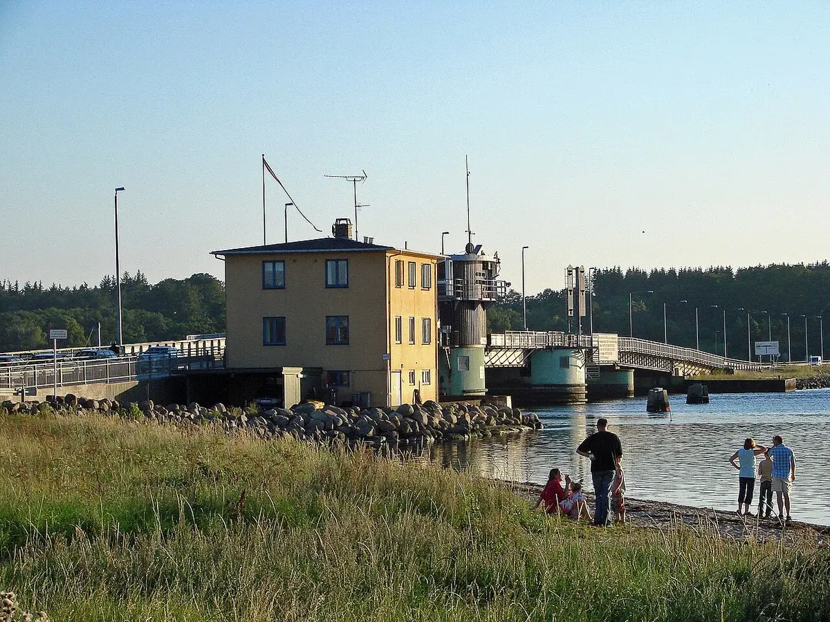 A bridge over water beside a small building, with people on the shore in the foreground.