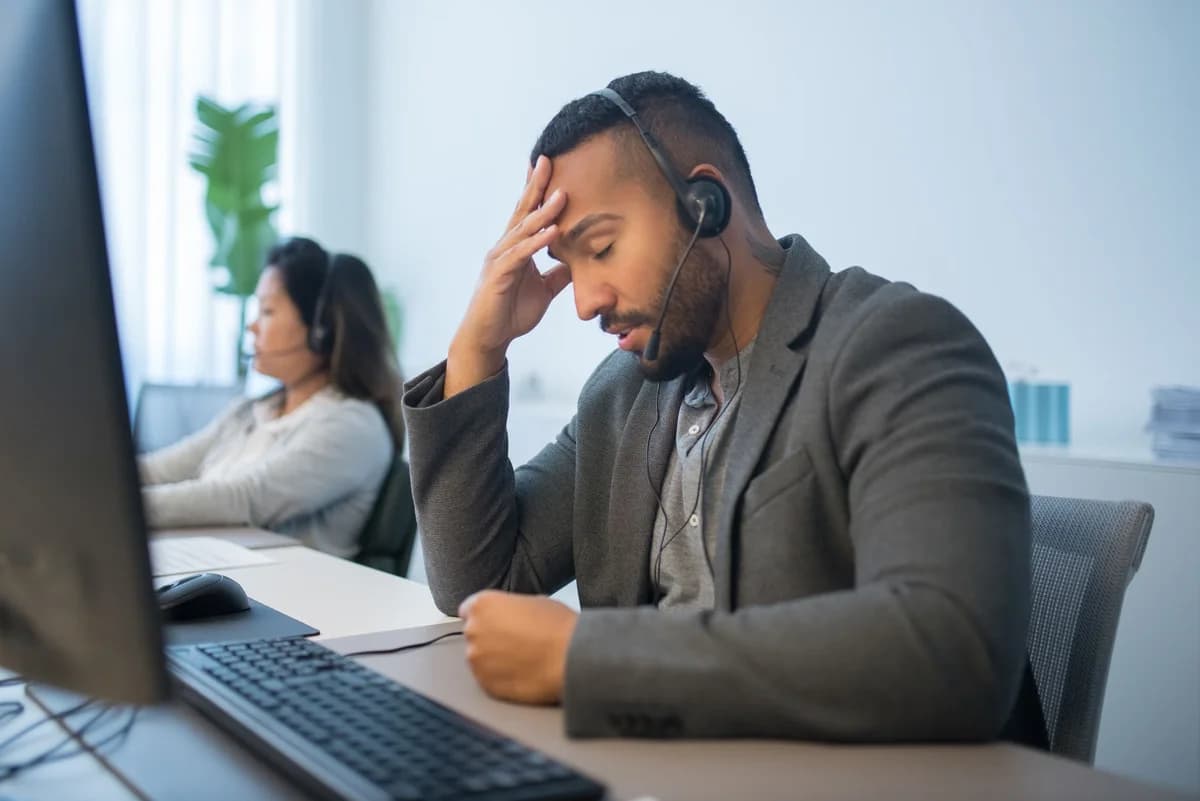 A stressed call center worker with a headset sits at his desk in an office.