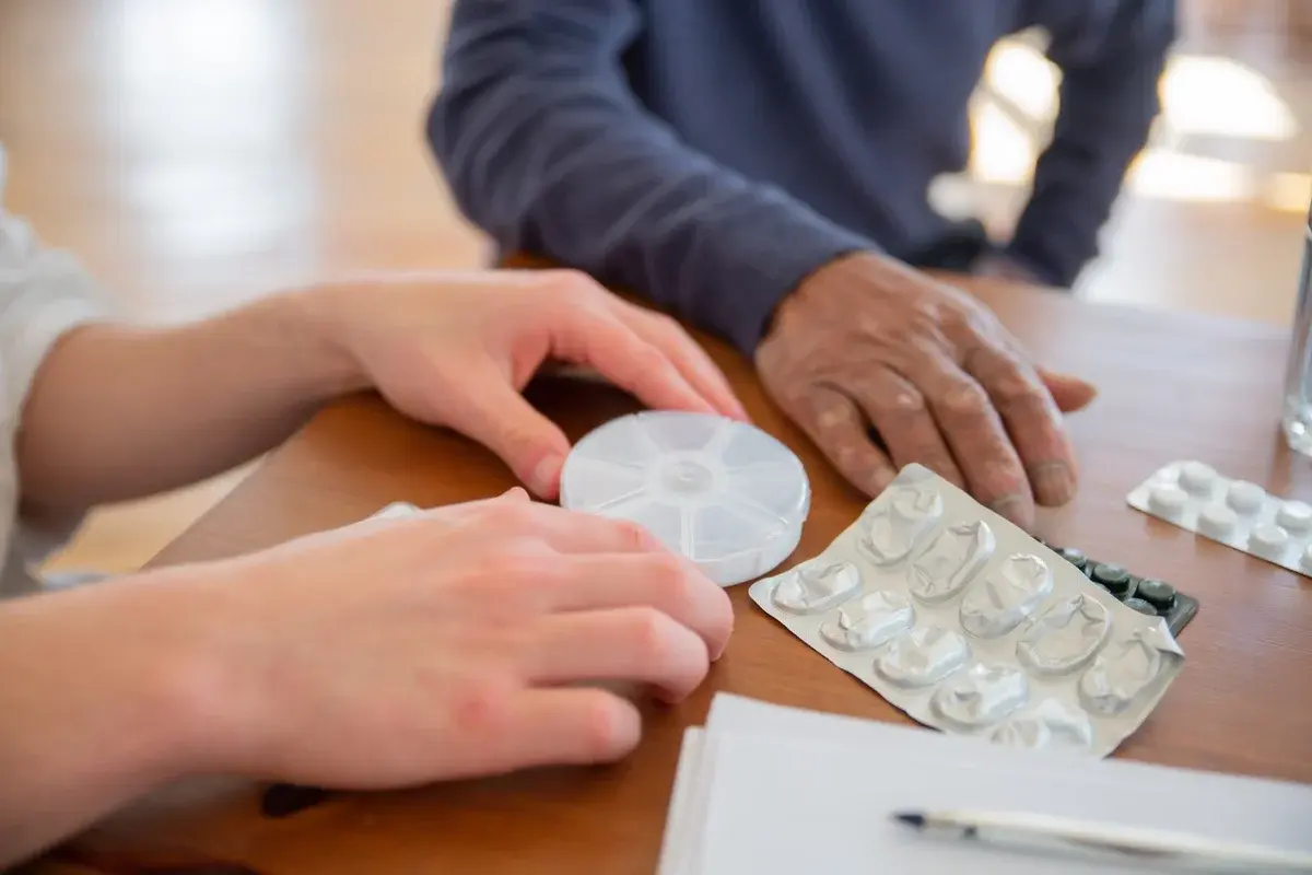 Hands sorting medicine beside a pill organizer on a wooden table.