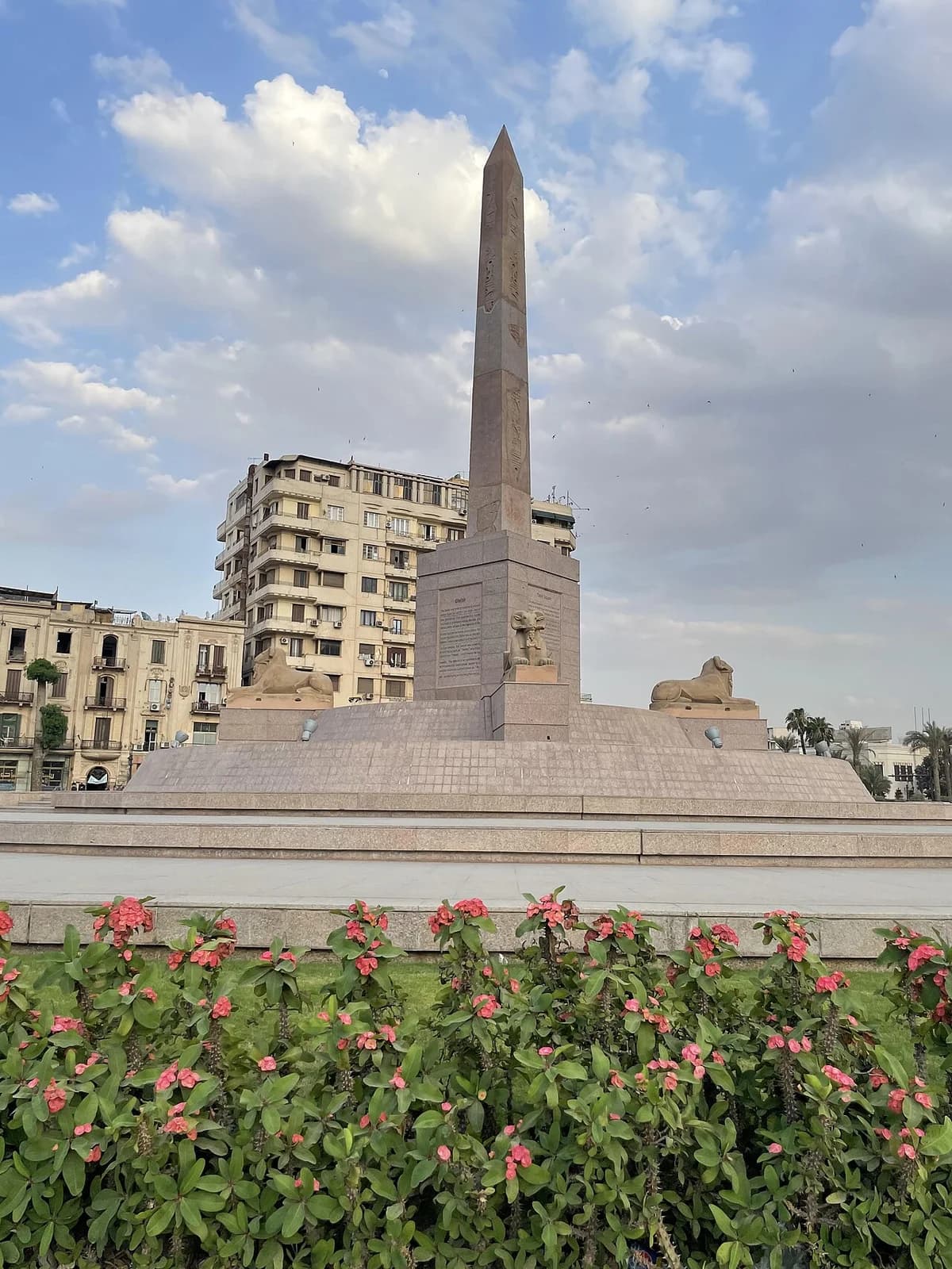 An obelisk monument in Tahrir Square, Cairo, with sphinx statues and city buildings behind it.