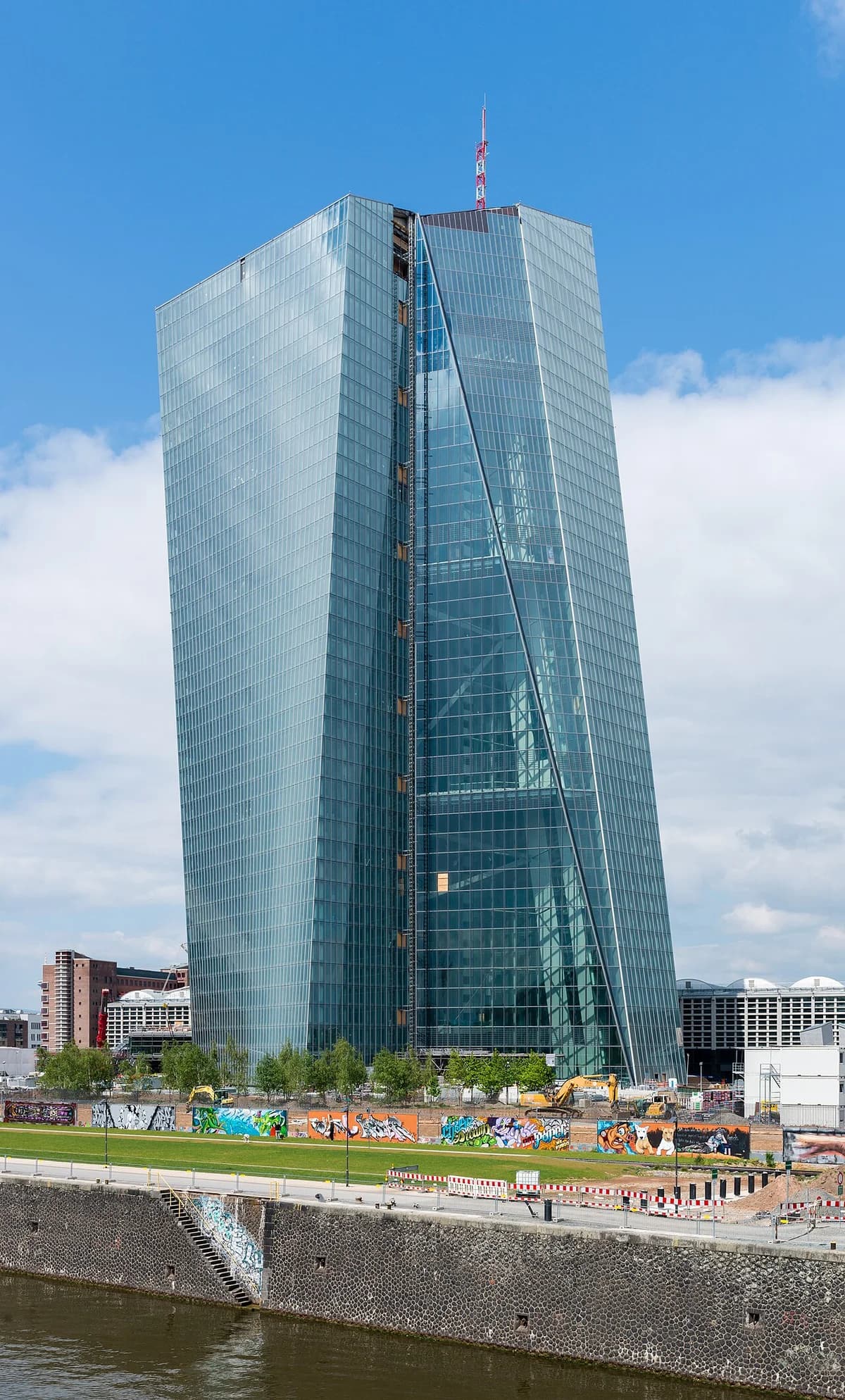European Central Bank tower in Frankfurt beside a construction site and riverfront wall.