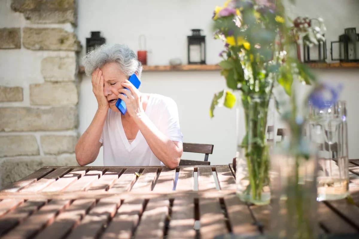 An elderly woman sits outdoors at a wooden table, holding a blue phone to her ear and covering her face in distress.