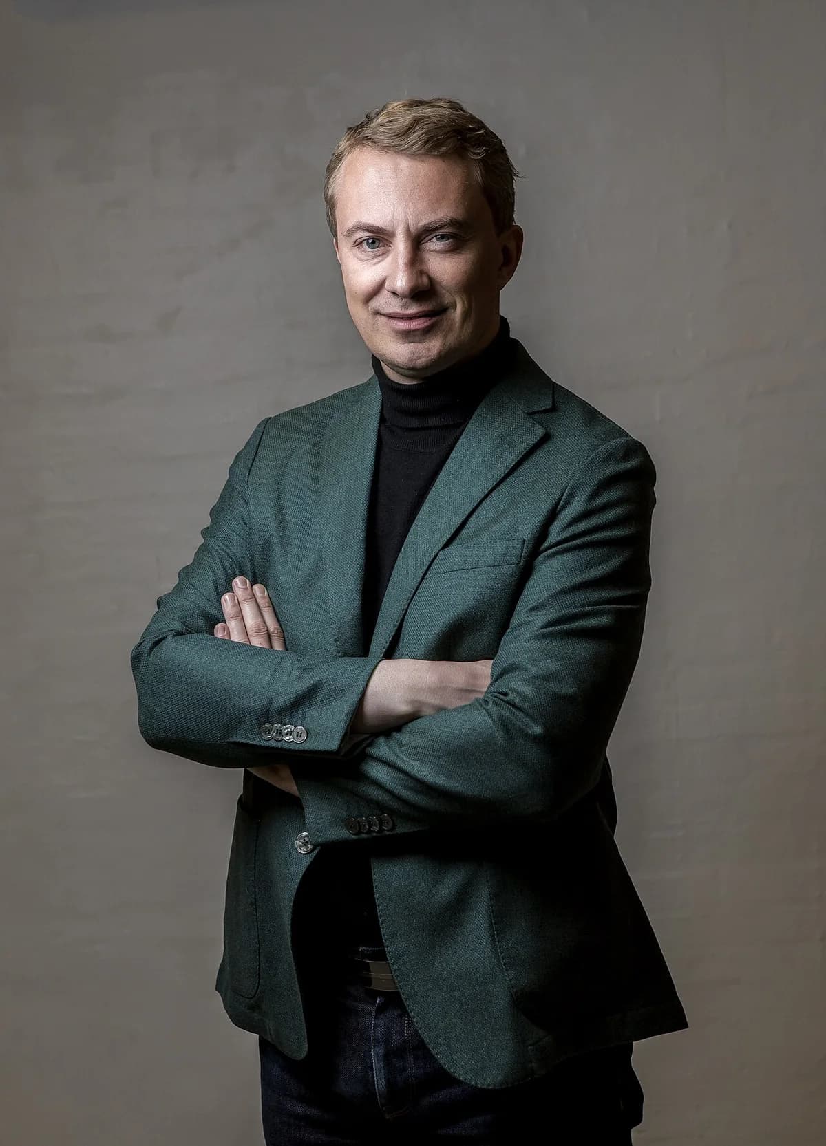 Studio portrait of a blond white man in a grey-green blazer and black turtleneck with arms crossed.