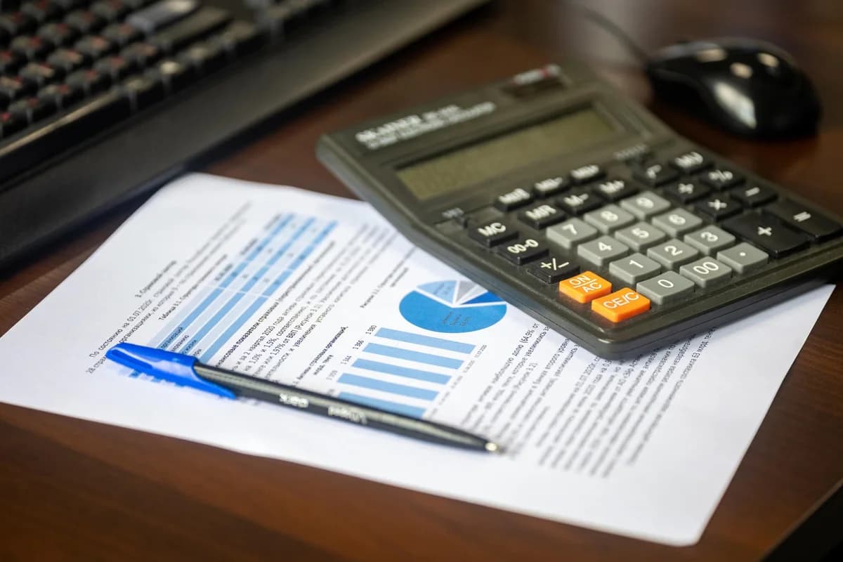 Calculator on top of a financial report with charts, alongside a pen, keyboard, and mouse on a desk.