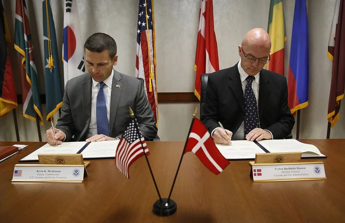 Two officials signing a cooperation memorandum with American and Danish flags on the desk