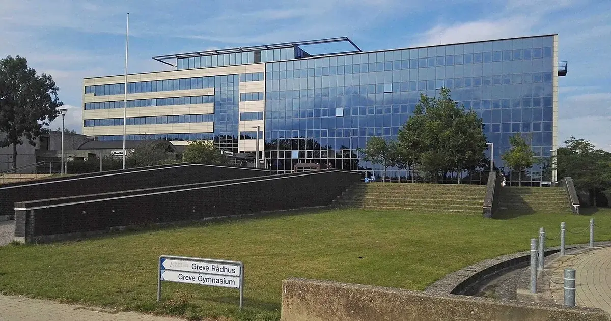 Modern glass town hall building with a grassy foreground and a sign for Greve Rådhus.