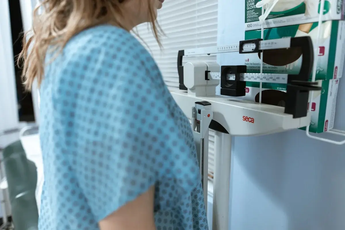 A woman in a clinic being weighed on a medical scale.