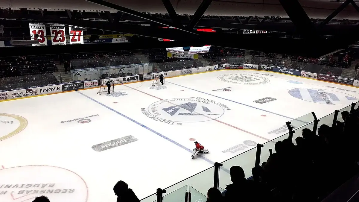 Indoor ice hockey rink with players on the ice and spectators in shadowed stands.