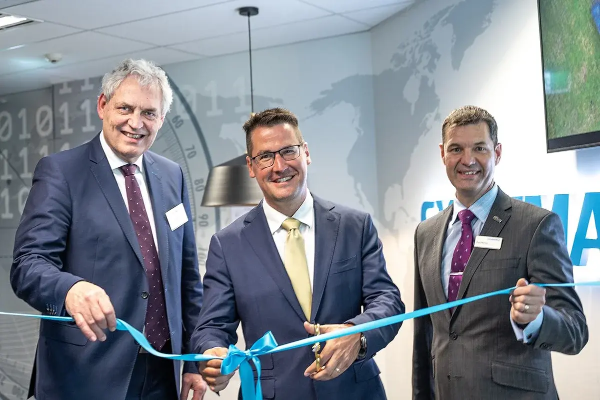 Three men in suits cutting a blue ribbon indoors at an event