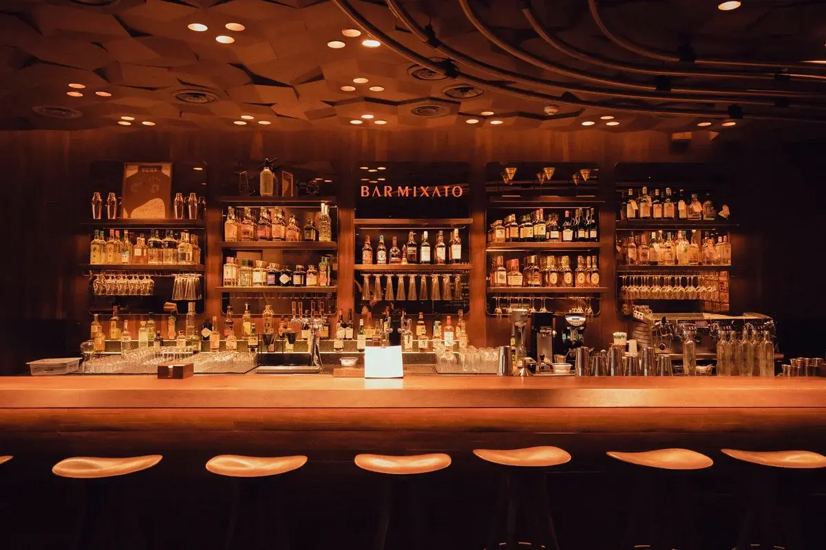 Warmly lit bar interior with wooden shelves and many bottles behind the counter.