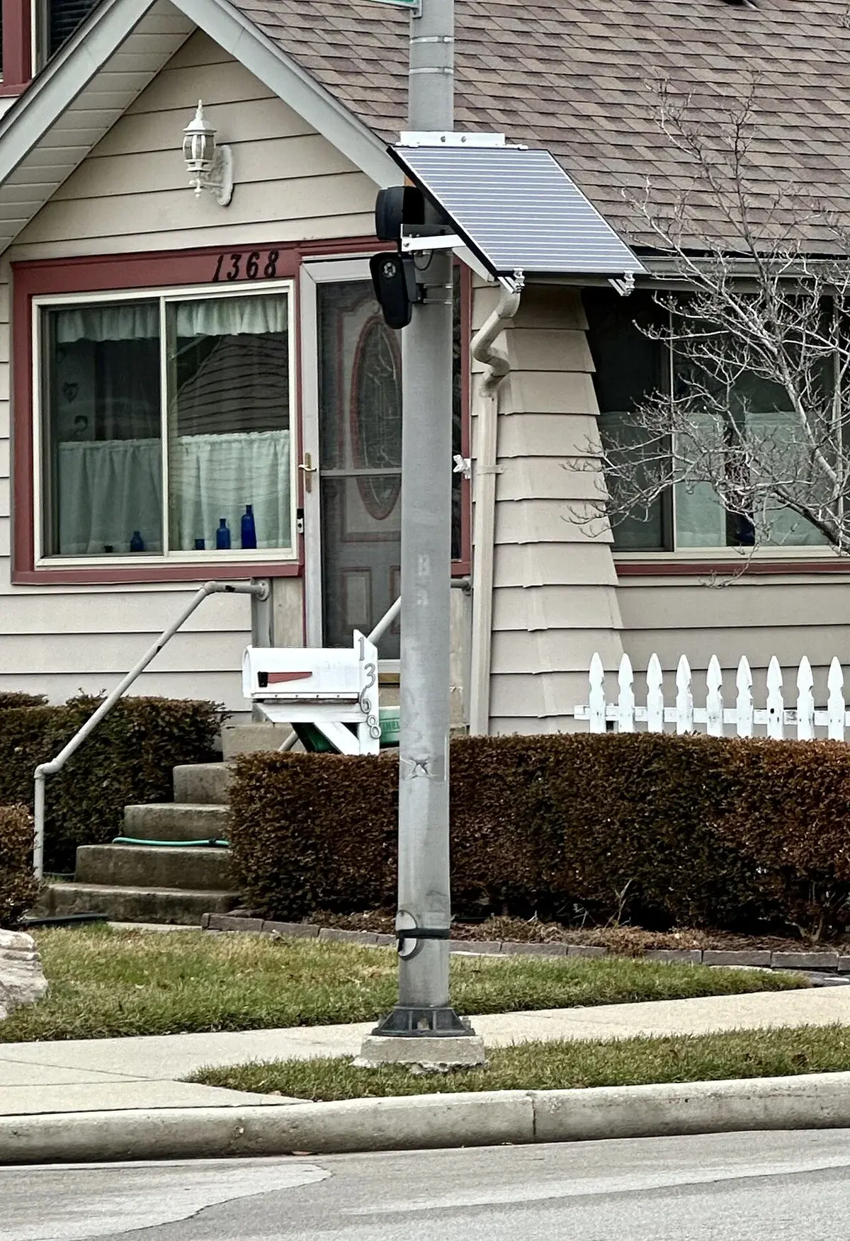A streetlight pole with a mounted security camera and solar panel outside a house.