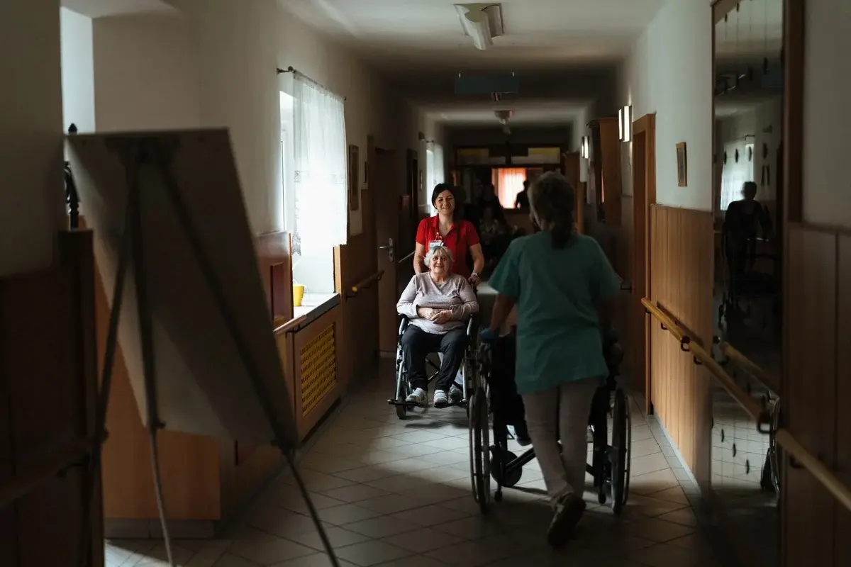 Caregiver pushing elderly woman in wheelchair through hallway
