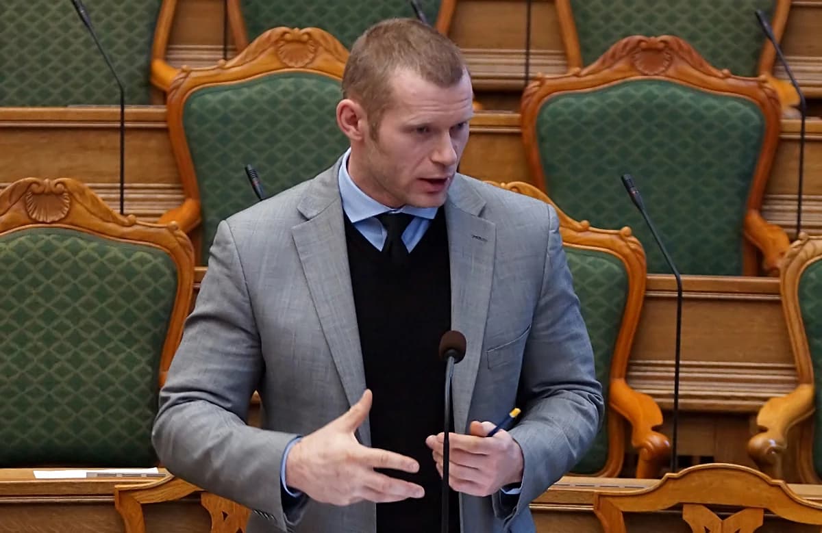 A man speaking at a podium in the Danish parliament chamber.