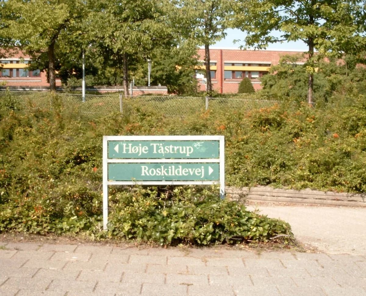 A green directional sign points to Høje Taastrup and Roskildevej beside a path with bushes and trees.