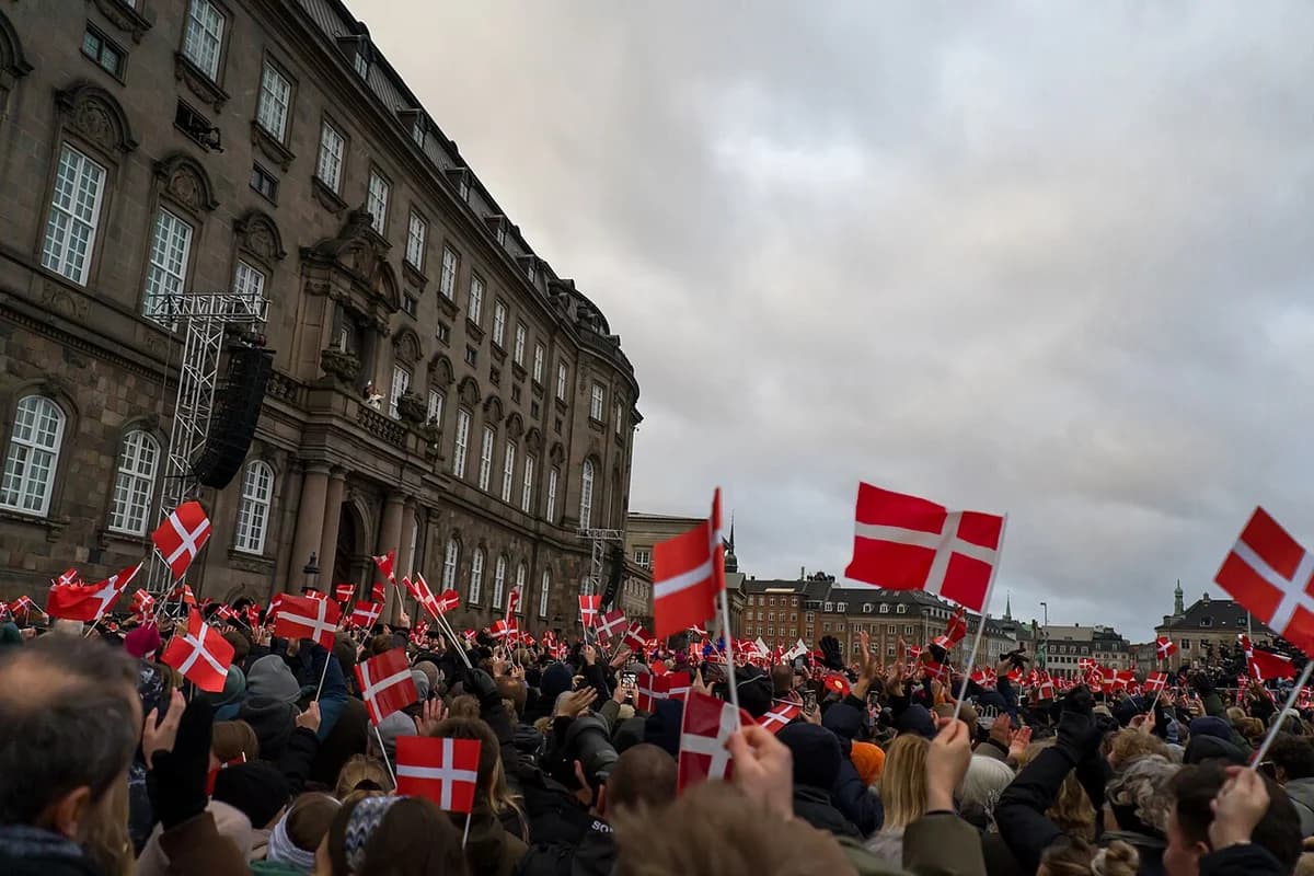 Crowd waving Danish flags outside a palace during a royal proclamation.