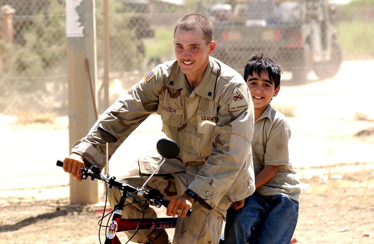 An American soldier rides a bicycle with an Iraqi boy sitting behind him.