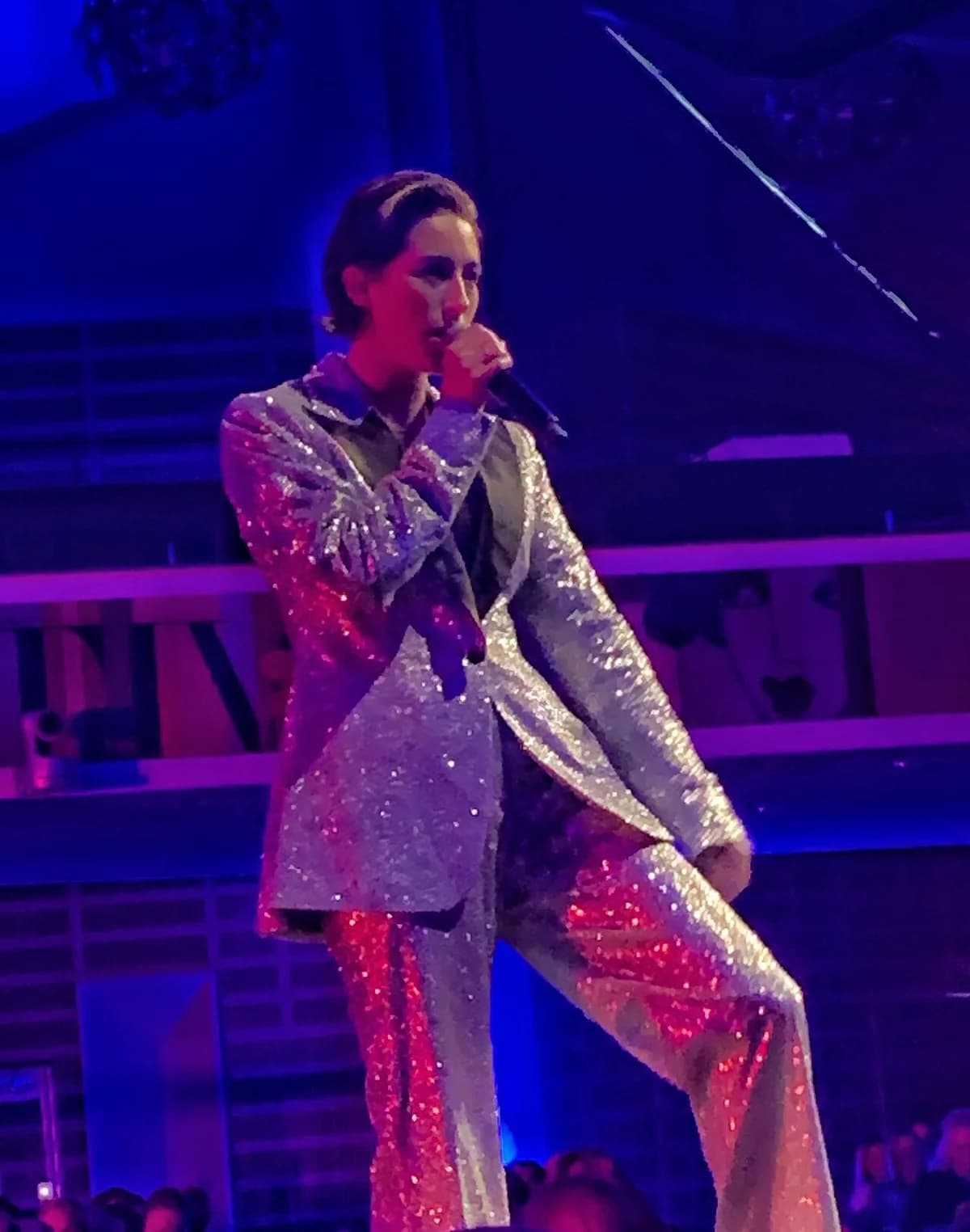 A singer performs on stage in a sparkling silver outfit under blue and purple lights.