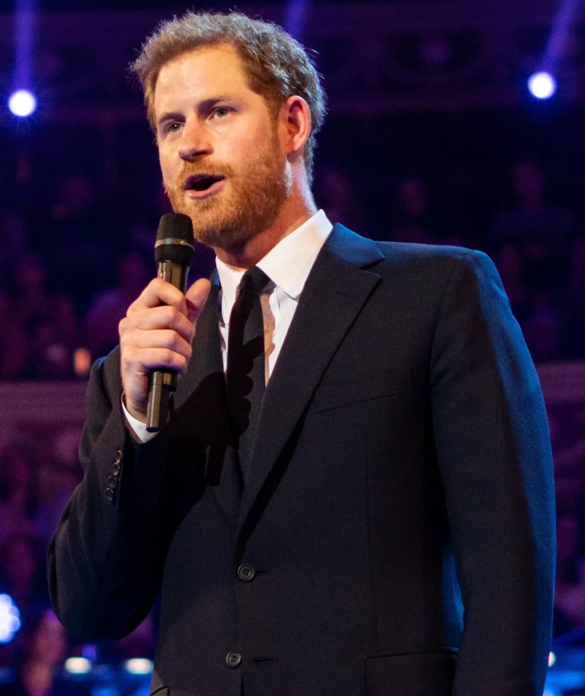 Prince Harry speaking into a microphone on stage in a dark suit.