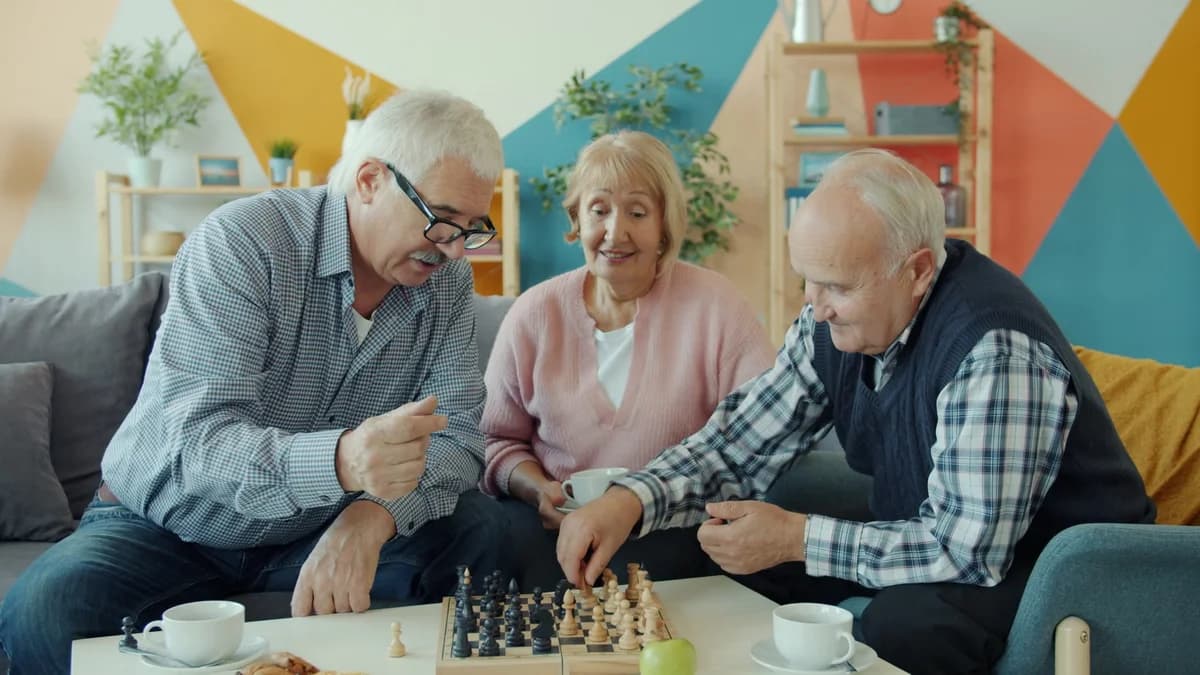Three seniors play chess together in a bright living room.